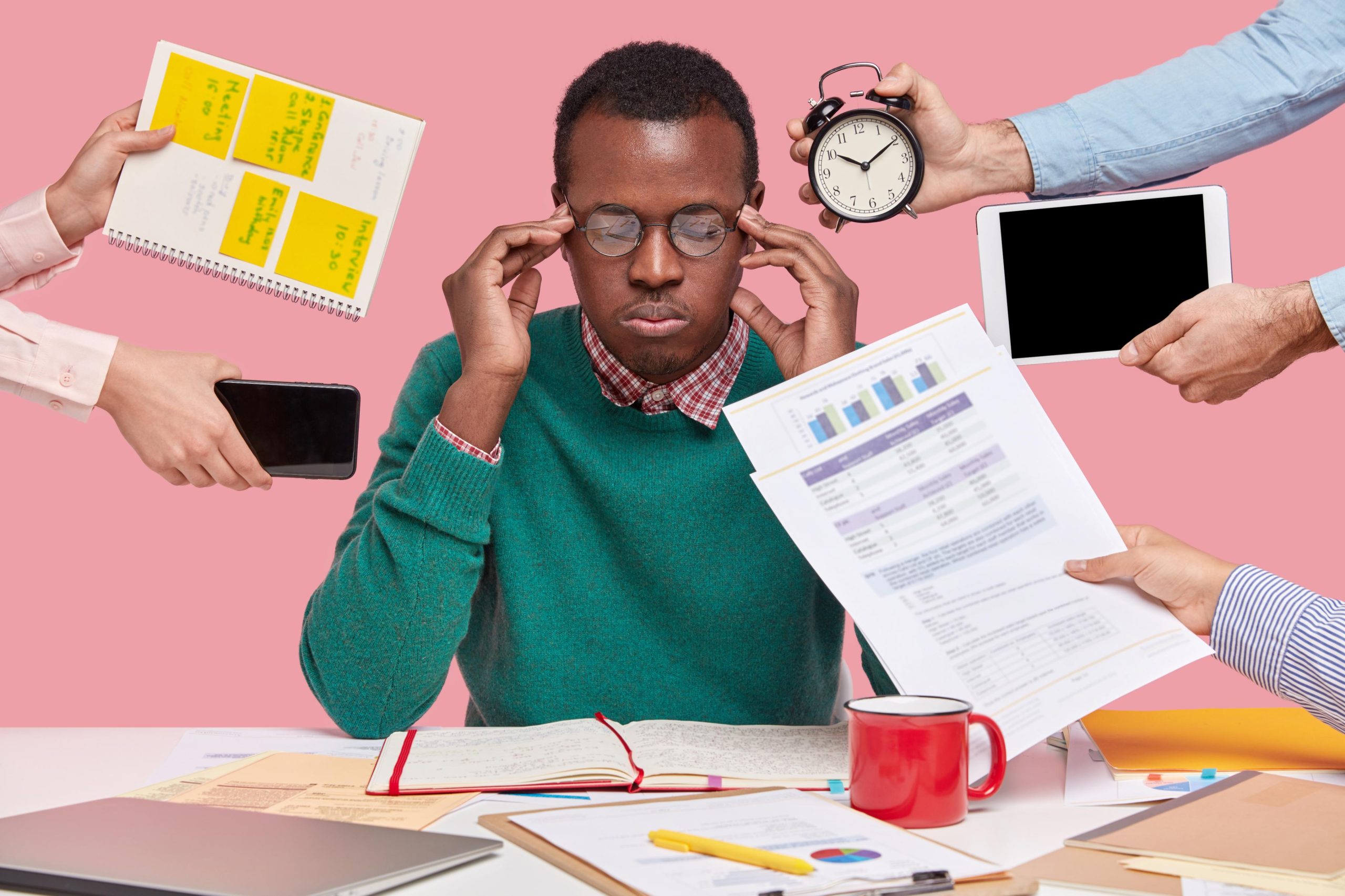 African-American man sitting at a desk surrounded by gadgets, papers, and a laptop, appearing focused.