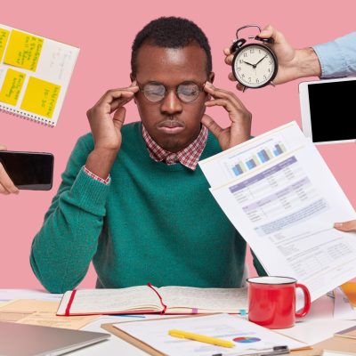 African-American man sitting at a desk surrounded by gadgets, papers, and a laptop, appearing focused.